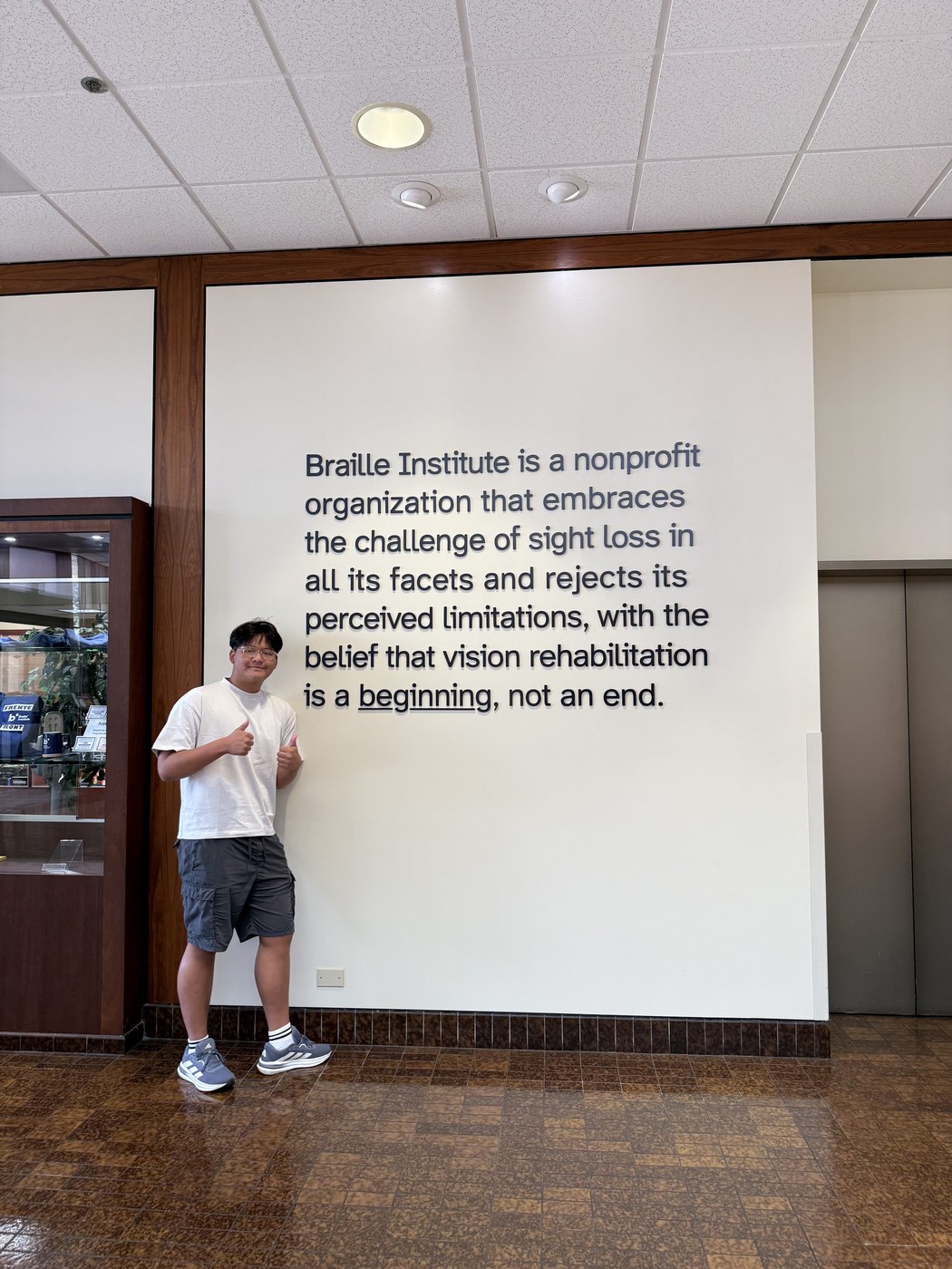 Ashton H. Jung next to a wall reading: Braille Institute is a nonprofit organization that embraces the challenge of sight loss in all its facets and rejects its perceived limitations, with the belief that vision rehabilitation is a beginning, not an end.