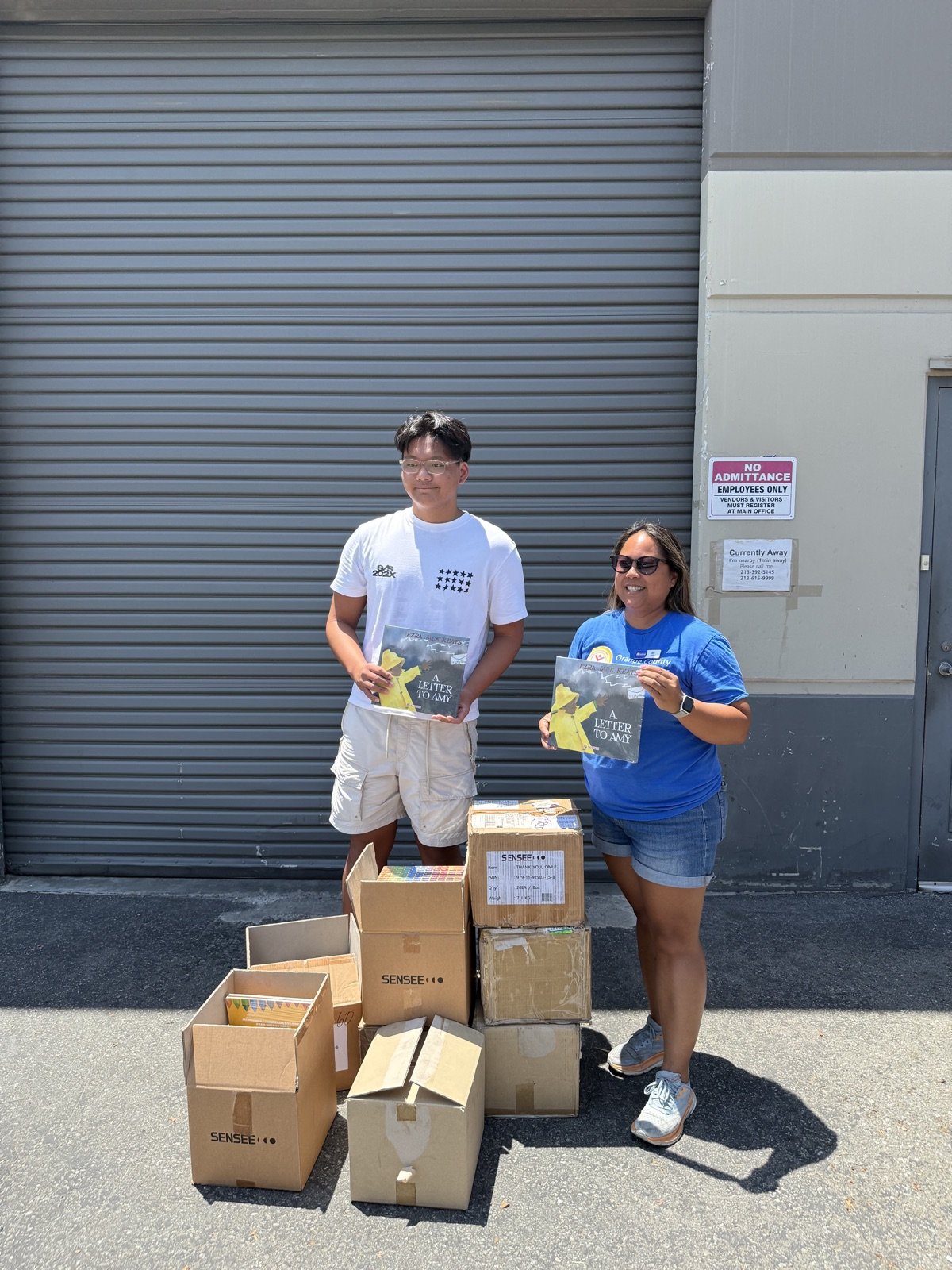 Ashton H. Jung and an Orange County United Way representative each holding a donated children's book, with stacked boxes of braille and tactile books between them